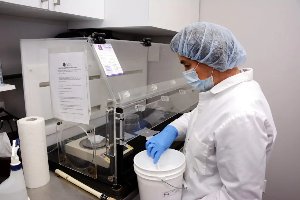 Masked lab technician scooping powder into a clear glovebox in a clean laboratory environment.
